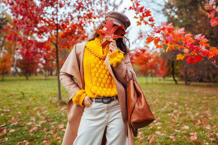 Stylish woman wears yellow sweater, coat holding handbag and red leaves walking in autumn park hiding face behind leaves. Fall female clothes and accessories. Fashionの写真素材