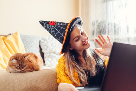 Halloween celebration at home. Happy smiling woman in witch hat waving hi online through video chat on laptop by her cat sleeping in couch. Restrictions due to covid pandemic.の写真素材
