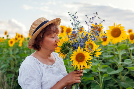 Senior woman in straw hat smells bouquet of yellow sunflowers in summer field at sunset. Middle-aged woman admires blooms relaxing outdoorsの写真素材
