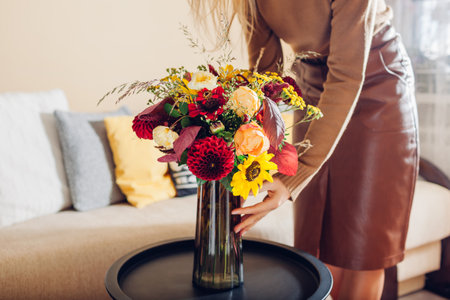 Woman puts vase with bouquet of sunflowers dahlias roses and zinnia flowers on table. Interior and fall decor at home.の写真素材