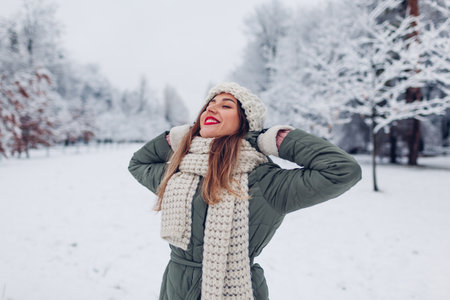 Portrait of happy young woman walking in snowy winter park wearing warm knitted clothes and red festive lipstick enjoying weather and landscape.の写真素材