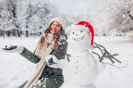 Happy woman tossing snow up by snowman in Santa hat playing outdoors in snowy winter park. Christmas festive season. Fun activitiesの写真素材