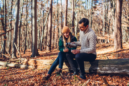 Happy loving couple drinking tea from flask relaxing in autumn forest. Man and woman sitting on trunk pouring coffee from vacuum flaskの写真素材