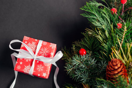 Top view of Christmas and New Year gift box wrapped in festive red paper and ribbon by fir wreath. Present for winter holidays on black background. Spaceの写真素材