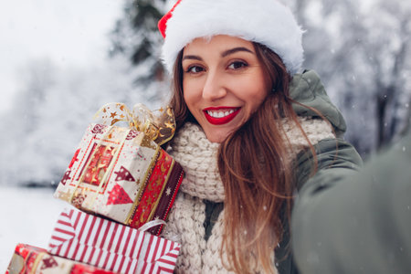 Portrait of woman taking selfie holding Christmas presents gift boxes in snowy winter park outdoors. Festive holiday season. Girl wearing Santa hat and red lipstickの写真素材
