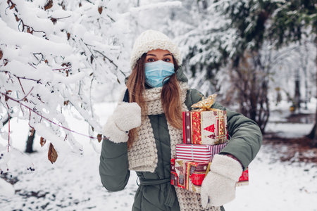 Woman holding Christmas presents gift boxes in snowy winter park wearing medical mask. Covid coronavirusの写真素材