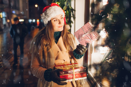 Portrait of young woman in Santas hat holding presents gift boxes by store showcase on city street at Christmas season.の写真素材