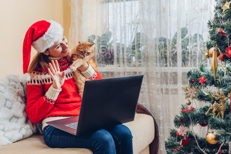 Christmas celebration with cat. Woman in Santas hat holding pet using laptop by New year tree at home.の写真素材