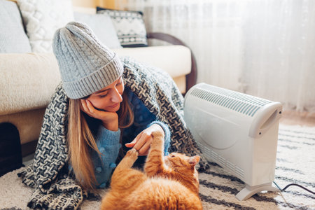 Using heater at home in winter. Woman playing with cat by device wearing sweater and hat under blanket. Heating season.の写真素材
