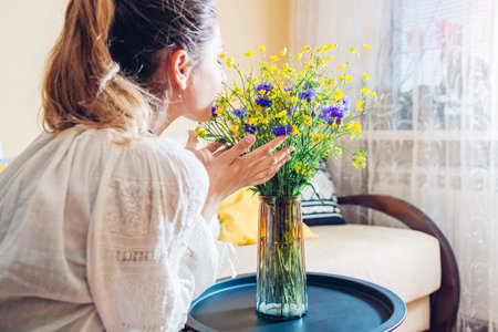 Woman in traditional shirt smells bouquet of blue and yellow flowers on table at home. Interior decor with Ukrainian colorsの写真素材