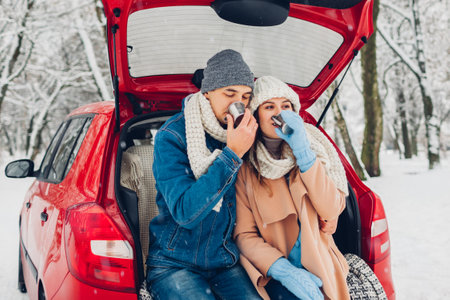 Valentine's day. Couple in love sitting in car trunk drinking hot tea in snowy winter forest. People relaxing outdoors during road trip. Having warm beverages enjoying landscapeの写真素材