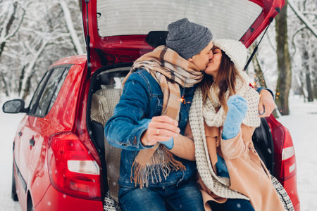Young couple in love kissing burning sparklers in car trunk in snowy winter forest. People celebrating Christmas outdoors during holiday road tripの写真素材