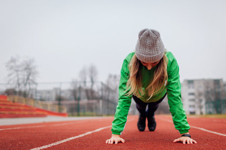 Young woman in sportswear doing plank or push ups outdoors on winter sportsground. Workout on track....