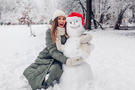 Happy young woman hugs snowman in snowy winter park dressed in Christmas Santa hat. Festive fun outdoor activities during cold weatherの写真素材