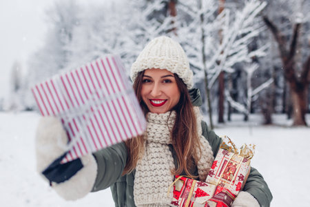 Happy woman holding Christmas presents gift boxes in snowy winter park offering one. Festive holiday season. Space. Girl wearing warm knitted clothes and red lipstickの写真素材