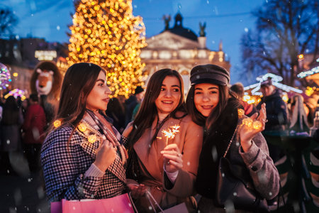 New Year eve celebration. Women friends burning sparklers in Lviv by Christmas tree on street fair celebrating holidays. Happy girls holding shopping bags under snow. Partyの写真素材