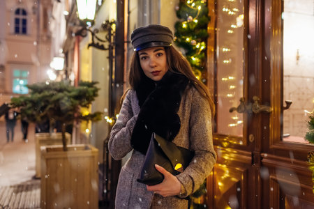 Christmas, New Year outdoor winter fashion. Woman walking on city street by decorated with lights showcases. Stylish girl holding purse under falling snowの写真素材