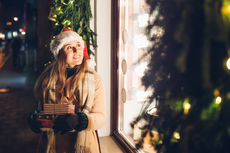 Excited young woman in Santa's hat holding presents gift boxes by store showcase on city street at Christmas season. Girl walking by decorated festive shops shopping for holidaysの写真素材