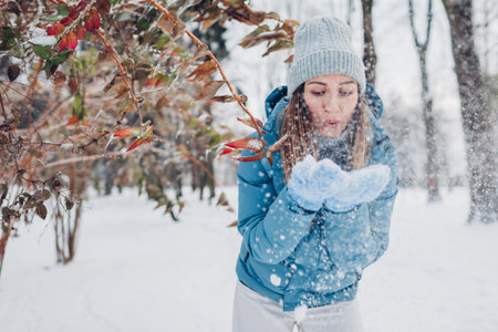Happy young woman blows snow from hands having fun in winter forest. Girl wearing blue coat and...