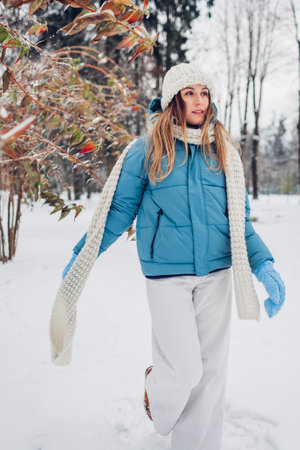 Full body portrait of young woman walking in snowy winter park wearing blue coat, knitted warm clothes. Female fashion for cold weatherの写真素材