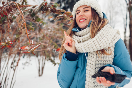 Portrait of happy woman listening to music on smartphone using headphones in snowy winter park. Girl dancing having fun outdoorsの写真素材