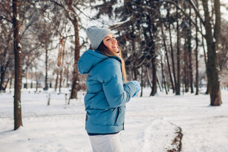 Happy young woman walking in snowy winter park wearing blue coat. Girl enjoys landscape wearing hat and mittens outdoors. Warm clothing for cold weatherの写真素材