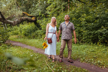 Stylish senior family couple walking outdoors in summer forest. Elderly people holding hands. Woman carries flowersの写真素材