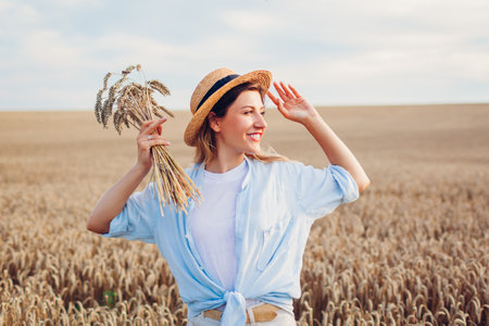 Portrait of smiling young woman walking among wheat in summer field wearing straw hat holding bundle of ripe wheatの写真素材