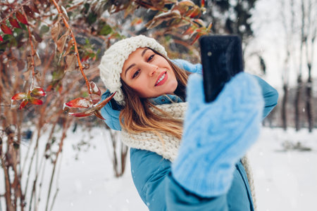 Happy woman taking selfie on smartphone in snowy winter park. Smiling girl takes pictures walking outdoors wearing warm clothing.の写真素材