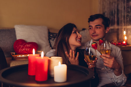 Valentines Day couple in love talking drinking wine with strawberries having romantic dinner celebrating at home. Celebrating with candles at nightの写真素材