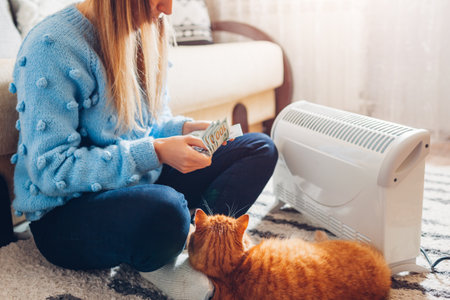 Expensive electricity. High prices for heating. Using heater at home in winter with cat. Sad woman counts money in cash wearing warm clothesの写真素材
