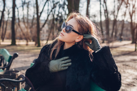 Close up portrait of stylish young woman wearing fur coat, green leather gloves and sunglasses in park. Spring trendy accessories. Girl relaxing in parkの写真素材