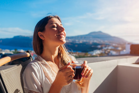 Woman relaxing on hotel terrace drinking morning coffee enjoying Santorini mountain landscape. Breakfast with viewの写真素材