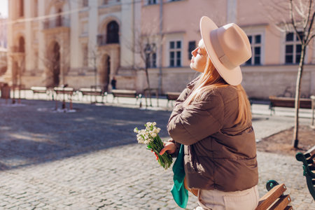 Back view of woman relaxing on bench outdoors on city street wearing stylish hat holding flowers. Spring fashionの写真素材
