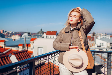Fashionable young woman enjoying landscape from balcony of European city holding hat. Spring fashionの写真素材
