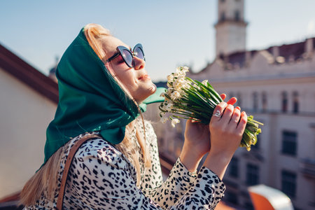 Stylish young woman wearing green retro shawl with sunglasses smelling spring flowers. Classic vintage outdoor fashionの写真素材