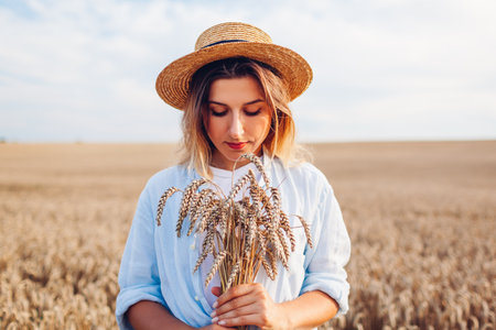 Portrait of young woman walking among wheat in summer field wearing straw hat holding bundle of ripe wheatの写真素材