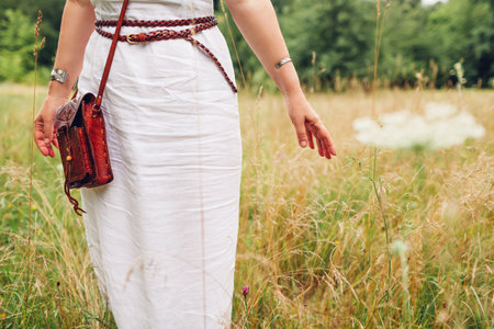 Boho style fashion. Woman wearing white dress, jewelry, carrying leather handmade handbag outdoors walking in meadowの写真素材