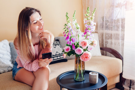 Woman relaxing using smartphone enjoying cozy interior with roses and foxgloves flowers in vase on table at home.の写真素材