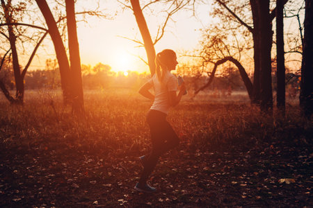 Young runner training in autumn park. Woman working out with water bottle at sunset. Active lifestyle. Silhouette of slim healthy girlの写真素材