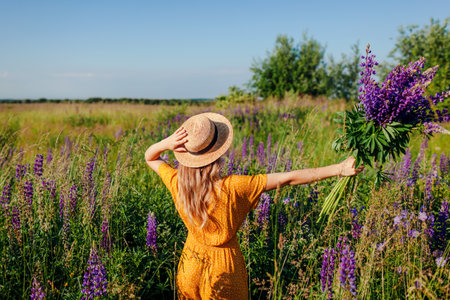 Happy woman holding bouquet of lupin flowers walking in summer meadow. Girl opens arms enjoying landscape and freedomの写真素材