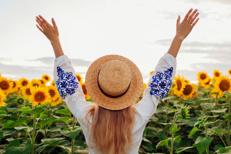 Back view of happy woman in hat and embroided dress raising arms in sunflower field at sunset. Peace and freedom in Ukraineの写真素材