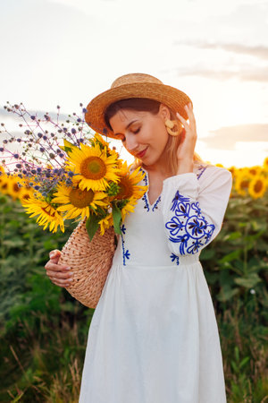 Young woman walking in blooming sunflower field at sunset smelling bouquet of flowers put in handbag. Girl wearing ethnic embroided clothingの写真素材