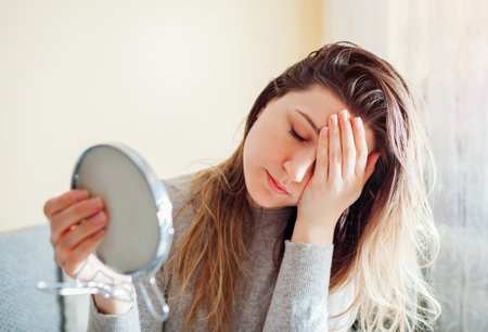 Stressed woman is sad about dirty oily and greasy hair looking in mirror at home. Healthcare. Hormonal imbalance issueの写真素材