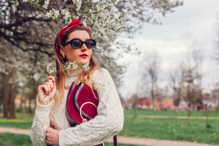 Portrait of woman wearing red hair scarf and sunglasses in spring park. Retro female fashion. Bright headscarf and purse. Stylish accessoriesの写真素材