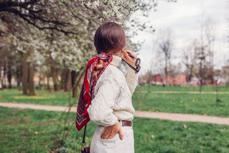 Back view portrait of stylish young woman wearing red hair scarf, white sweater, pants, sunglasses in spring park. Classic female fashion. Headscarf bandana for bun hairstyle. Bright accent accessoryの写真素材