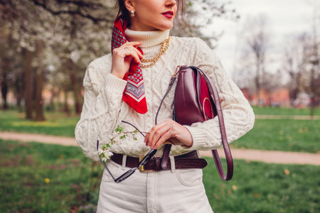 Close up portrait of woman wearing red hair scarf holding purse and sunglasses in spring park. Female fashion. Bright headscarf accessory with white clothesの写真素材