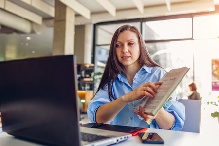 Young female teacher working through videoconference in office using laptop. Woman showing students book online. Remote lessonの写真素材