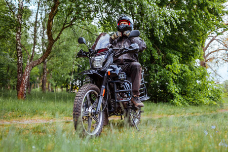 Biker unfastens helmet after riding motobike in summer forest. Man wearing leather clothes outdoors....