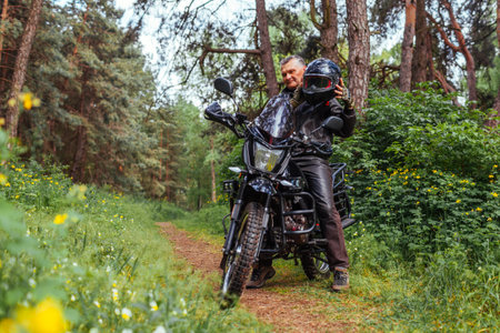 Senior biker taking helmet off after riding motobike in summer forest. Retired man wearing leather clothes sitting on motorcycle. Outdoor hobbyの写真素材